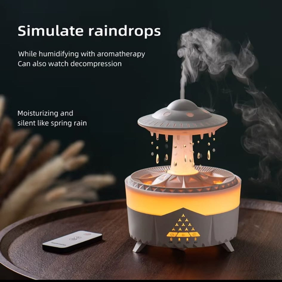A decorative rain simulator humidifier and diffuser shown on a wooden surface with steam rising from the top. The device has a gray upper dome, a glowing orange-yellow diffuser base, and small legs.